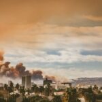 A city skyline with palm trees in the foreground, backed by mountains and dark smoke rising beneath a dramatic, orange-tinted sky.
