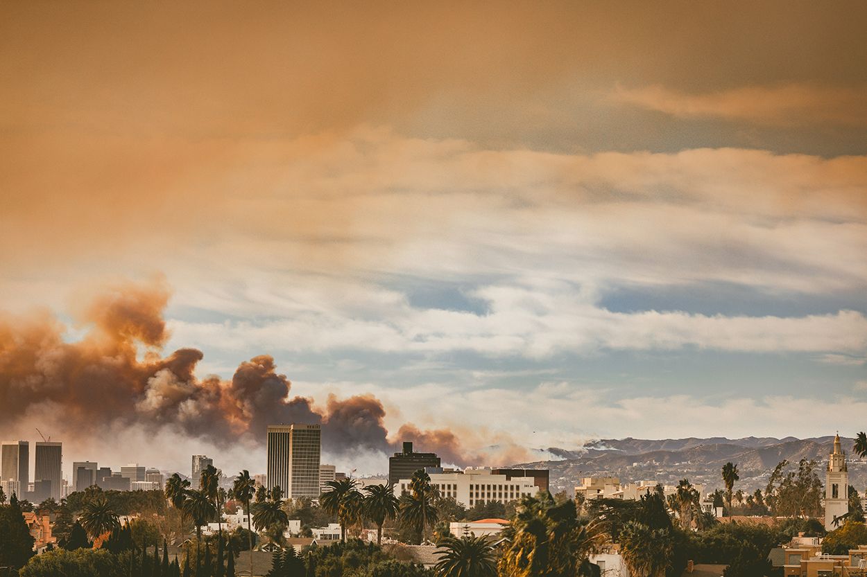 A city skyline with palm trees in the foreground, backed by mountains and dark smoke rising beneath a dramatic, orange-tinted sky.