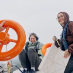 Lena Sunchugashevam, left, and Aisha Mardini, wearing life jackets, smile aboard an inflatable boat while another person holds an orange life ring in the foreground under an overcast sky on calm water.