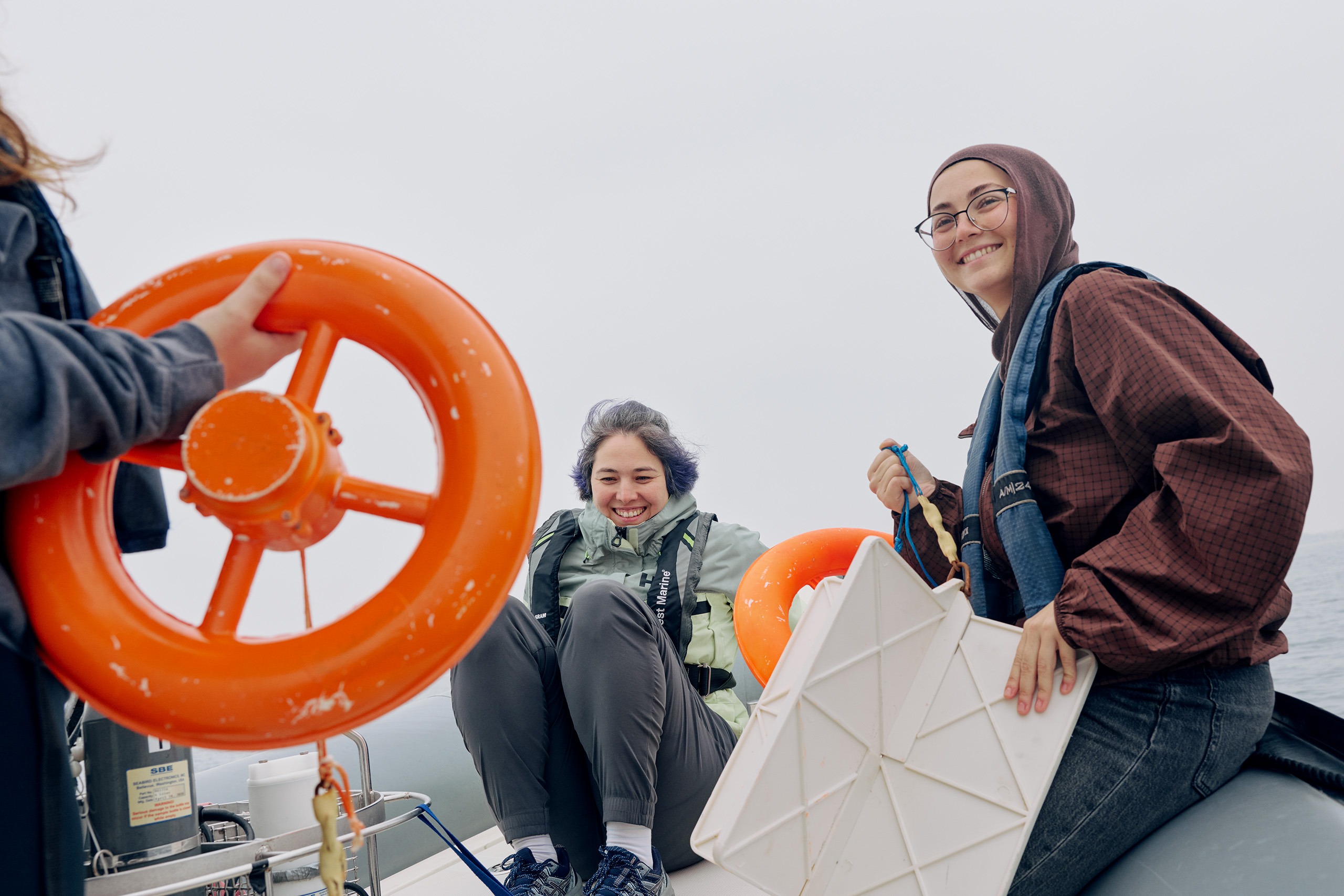 Lena Sunchugashevam, left, and Aisha Mardini, wearing life jackets, smile aboard an inflatable boat while another person holds an orange life ring in the foreground under an overcast sky on calm water.